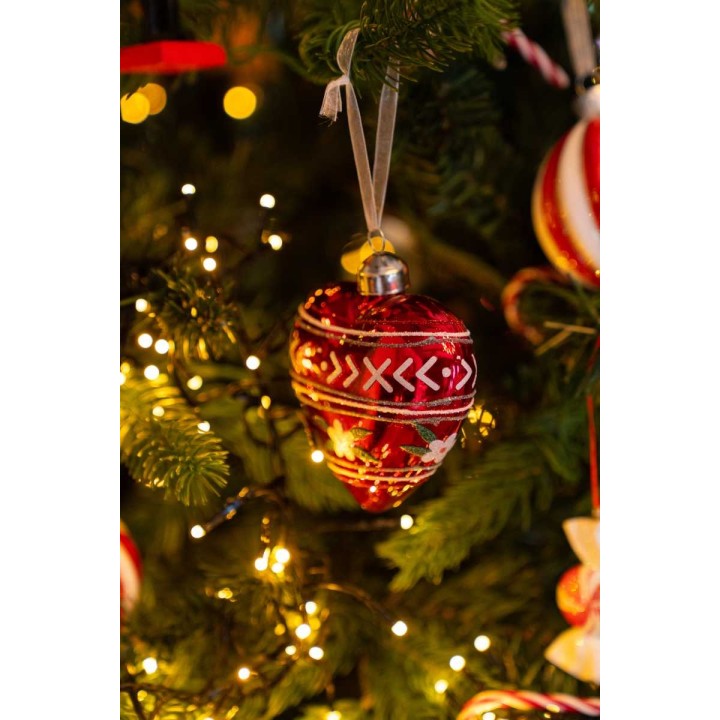 Een rode kerstbal met witte patronen, glanzend, hangend tussen groene takken en gouden lichtjes.