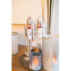 Two silver nutcracker figurines stand beside elegant candles on a warm wooden surface.
