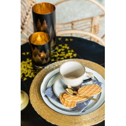 A cozy table setting with a golden woven placemat, a cup, and cookies beside elegant candles.