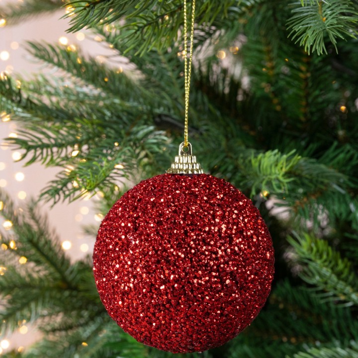 Glinsterende, rode kerstbal hangt aan een groene dennenboom met zachte achtergrondverlichting.