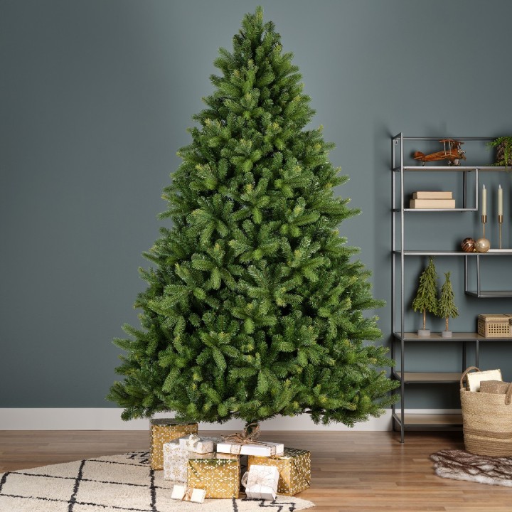 Een groene kunstkerstboom staat tegen een blauwe muur, omringd door cadeaus op een vloerkleed.