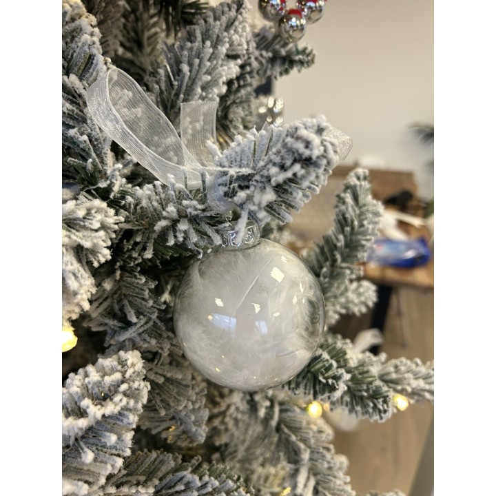 A frosted glass ornament hangs from a snowy green tree, adorned with a delicate ribbon.