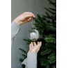 Een hand houdt een glazen kerstbal boven een andere hand, met een dennenboom op de achtergrond.