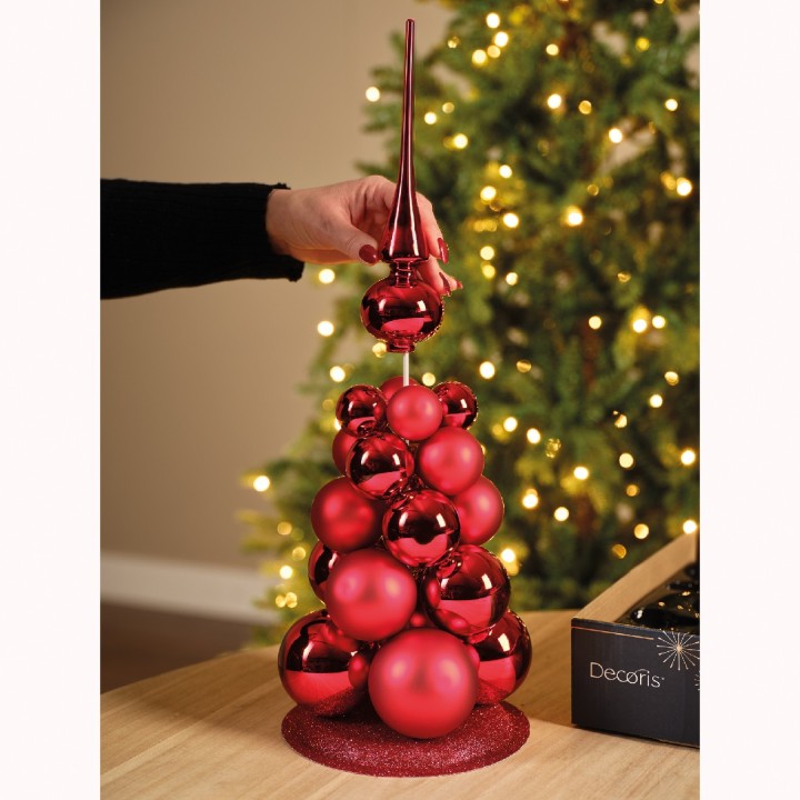 Een kerstboom van glanzende rode ballen met een sprankelende piek, op een houten tafel.