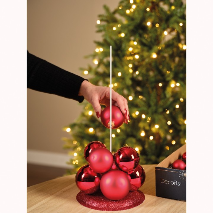 Een hand plaatst een glanzende rode kerstbal op een stapel, met een kerstboom op de achtergrond.