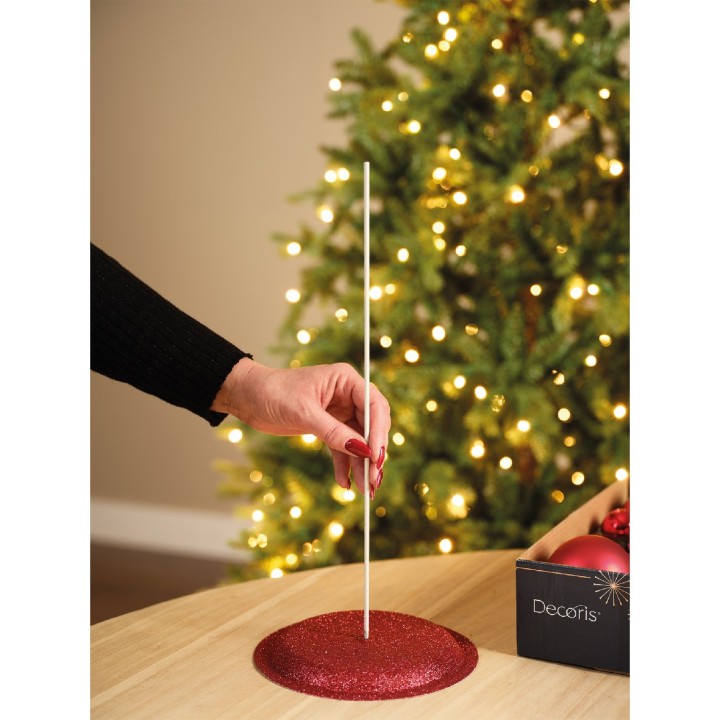 Hand plaatst een witte stok in een glinsterende rode houder, met een kerstboom op de achtergrond.