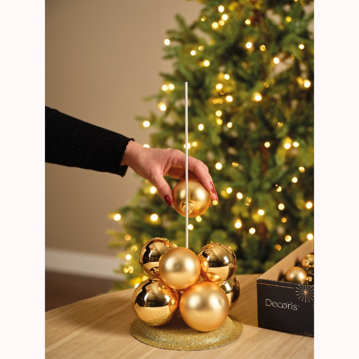 Een hand plaatst een gouden kerstbal op een stapel, omringd door lichtjes en een kerstboom.