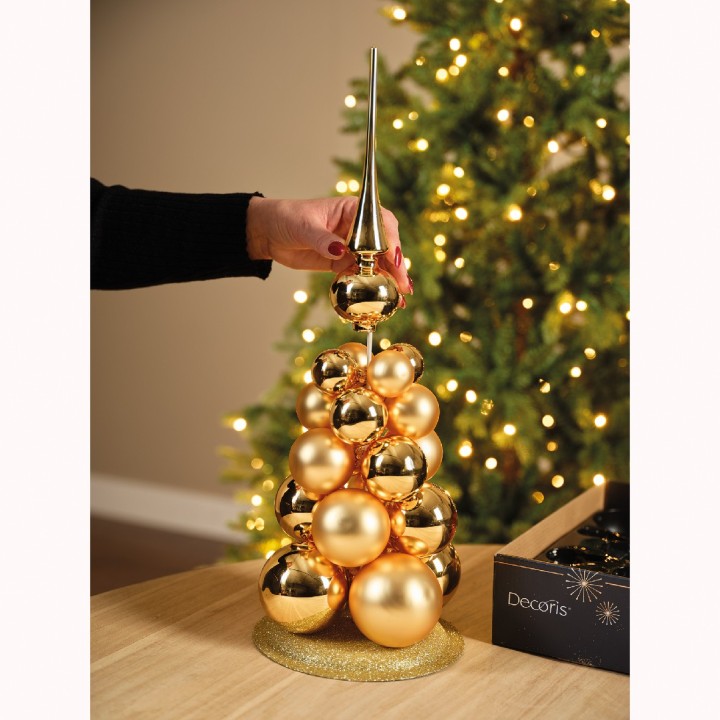Gouden kerstboom van glanzende ballen met een puntige top, sfeervol verlicht op een tafel.