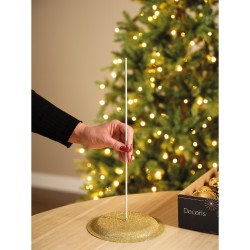 A hand inserts a thin white pole into a glittery gold base near a softly lit Christmas tree.