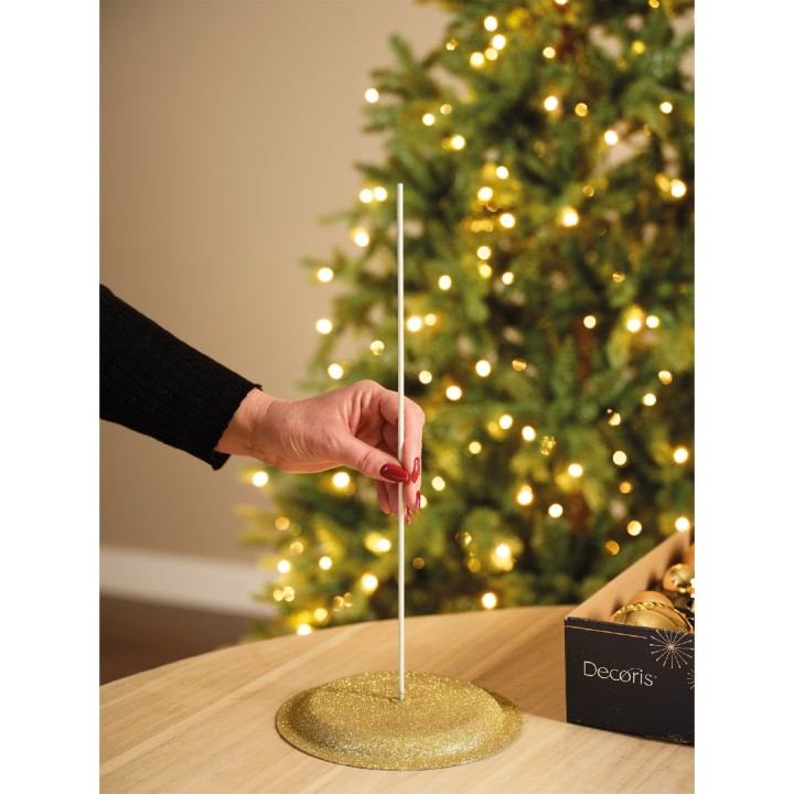 Een hand plaatst een witte stok in een gouden, glinsterende basis naast een kerstboom met lichtjes.